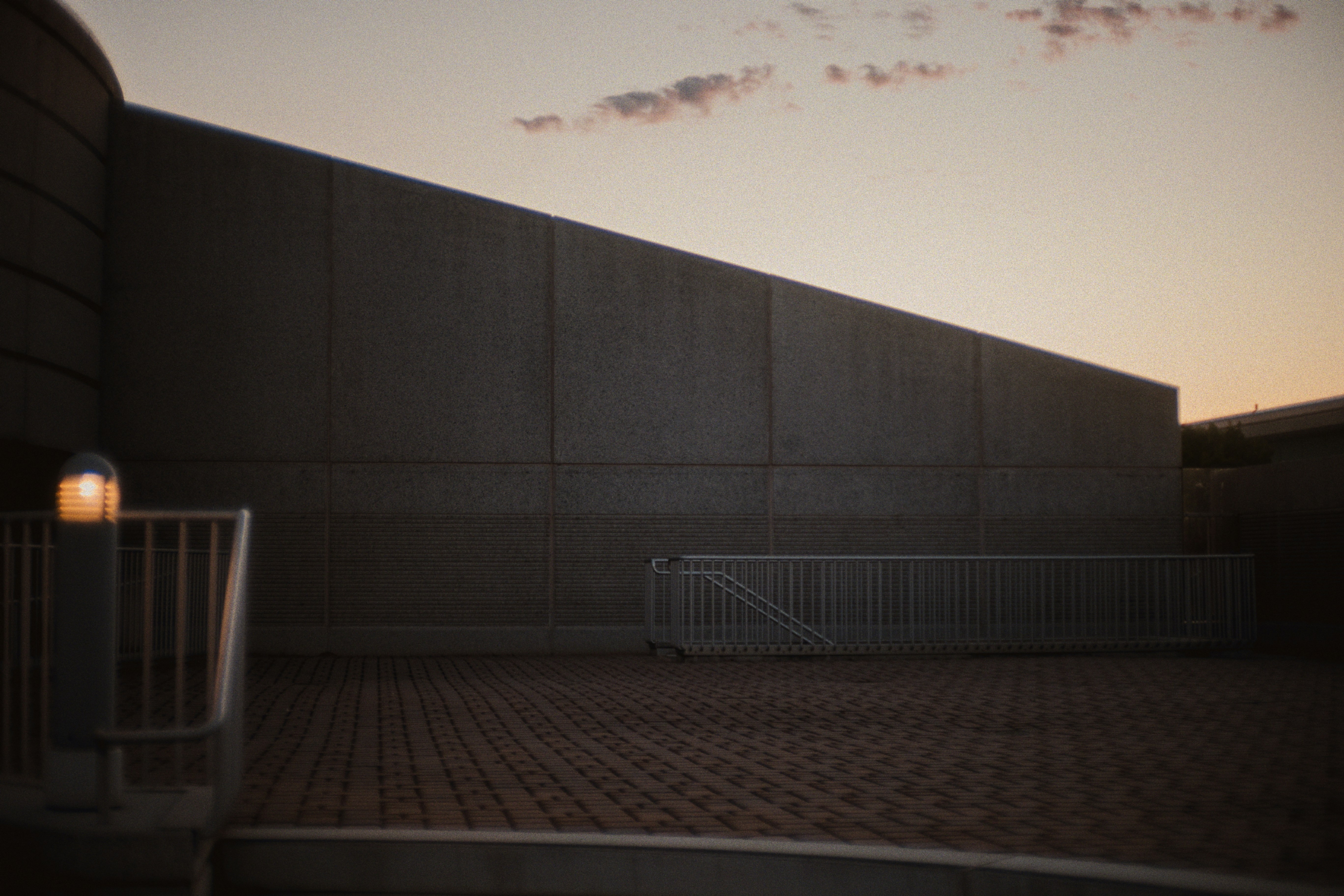 Minimalist concrete architecture at dusk: sloped textured wall, paved plaza, and a single warm bollard light.